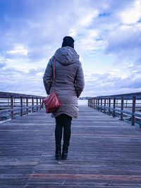 Rear view of woman standing in warm clothing on pier against cloudy sky