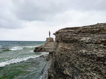 Mid distance view of man standing on cliff by sea against cloudy sky at byrums raukar