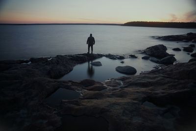 Rear view of man standing on rock at beach against sky during sunset