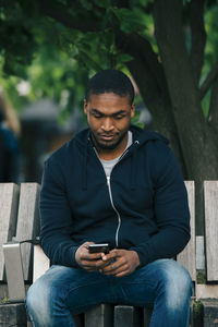 Young man using smart phone while sitting on bench in city