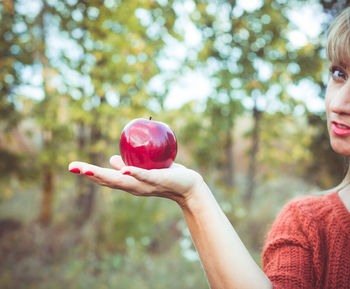 Cropped portrait of woman holding apple against trees