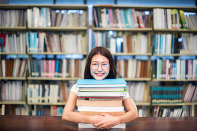 Portrait of smiling female student with stack books on table in library