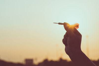 Cropped hand of person holding dart against sky during sunset