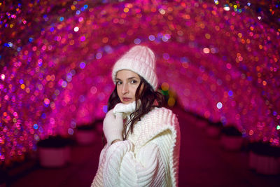 Woman in a white sweater, hat and white scarf on a background of pink lights