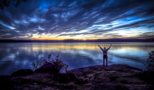 Man with arms raised standing at lakeshore against sky during sunset