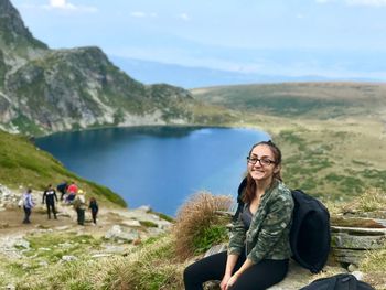 Portrait of smiling young woman sitting on by lake against sky