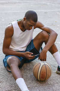 Sportsman with basketball using smart phone while sitting at court
