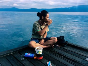 Young woman sitting by lake against sky