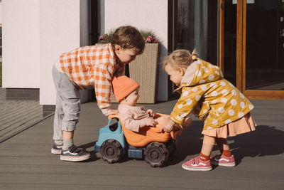 Brother sister giving a ride to baby sister in big toy car outdoors. children having fun near house