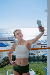 Happy woman in swimsuit taking selfie on mobile phone standing on the balcony of the hotel room