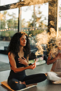 Woman with hand on chest holding burning sandalwood while sitting cross-legged at retreat center