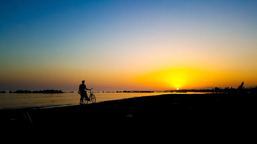 Silhouette man riding bicycle on beach against clear sky during sunset