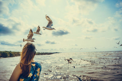 Rear view of woman looking at seagulls flying over sea against cloudy sky