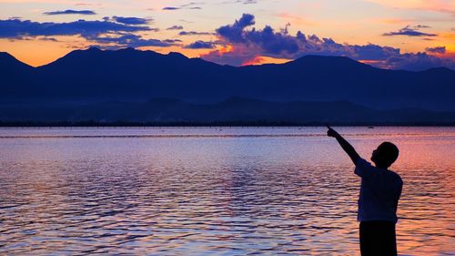 Boy pointing while standing by lake against sky during sunset