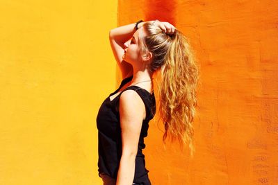 Side view of young woman with hand in hair standing against orange wall