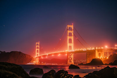 Illuminated suspension bridge over sea against sky during sunset