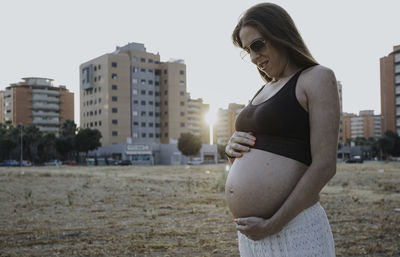 Smiling pregnant woman touching abdomen while standing against buildings and sky