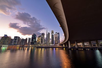 Illuminated modern buildings by river against sky during sunset