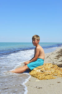 A boy sits on a yellow stone on the seashore. he has a white-toothed smile 