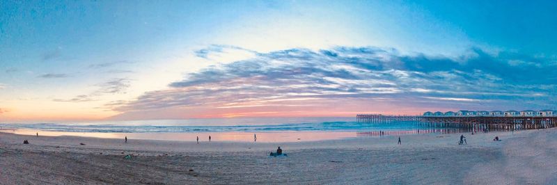 Panoramic view of people at beach against cloudy sky during sunset