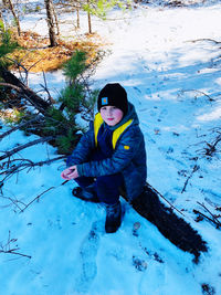 Full length portrait of boy sitting on fallen tree trunk during winter