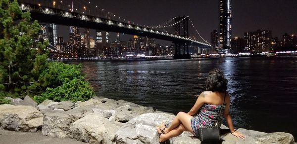 Rear view of woman sitting on rock against illuminated manhattan bridge