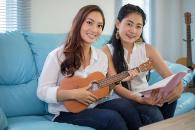 Portrait of smiling female friends with guitar sitting on sofa at home