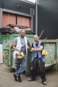 Full length of male construction workers standing with hardhat against metal container at site