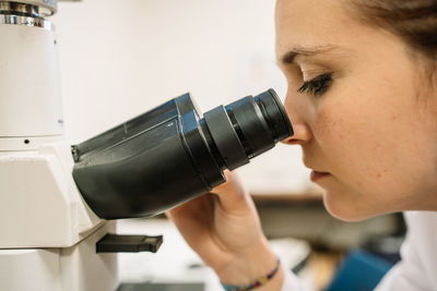 Side view of concentrated female scientist in white medical uniform looking through modern microscope while examining samples in light laboratory