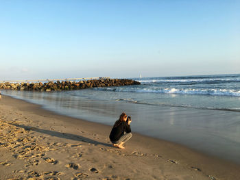 Rear view of woman crouching while photographing at beach