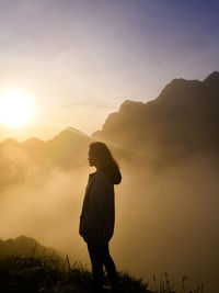 Side view of woman standing on mountain during foggy weather at sunrise