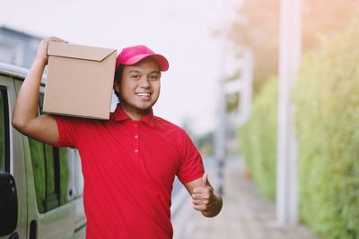 Portrait of smiling delivery person wearing cap standing with cardboard outdoors