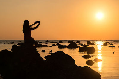Silhouette woman photographing while sitting on rock at beach against sky during sunset