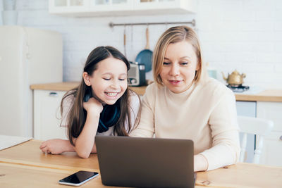 Smiling girl looking at mother using laptop on table in kitchen