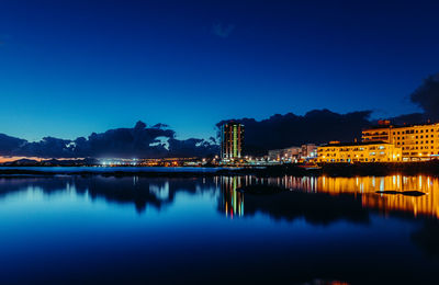Illuminated buildings by lake against blue sky at night