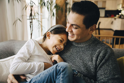 Smiling father and daughter spending leisure time while sitting on sofa at home