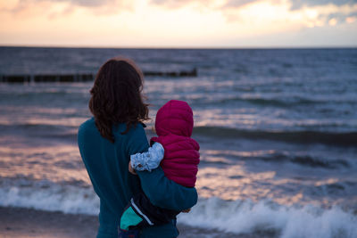 Rear view of mother with baby standing on shore at beach during sunset