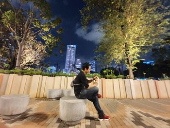 Side view of man sitting with bottle on seat in city at night