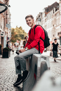 Smiling man with backpack sitting on barricade amidst street in city