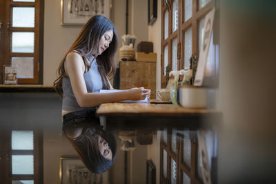 Side view of young woman reading book while sitting on table at home