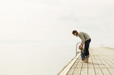 Full length side view of mother and baby girl standing on pier at sea against sky