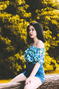 Portrait of beautiful young woman sitting on log against trees at park