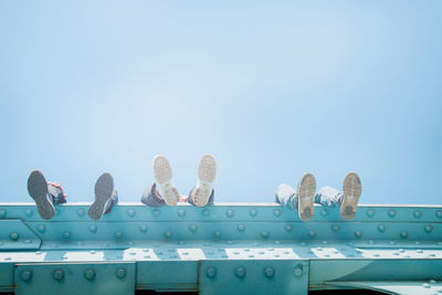 Low section of people wearing shoes while sitting on bridge against clear sky during sunny day