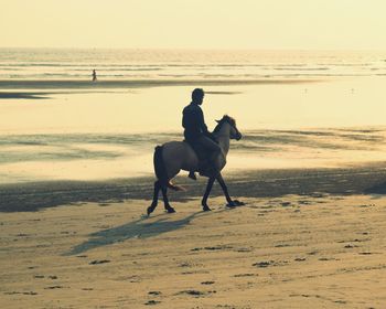 Man riding horse on beach against sky during sunset