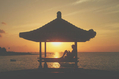 Silhouette man sitting by sea against sky during sunset