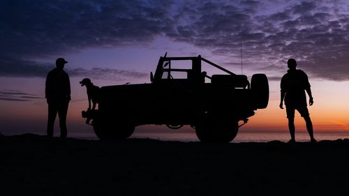 Silhouette people standing with dog and vehicle against sky during sunset