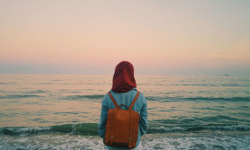 Rear view of woman carrying backpack standing at sea shore during sunset