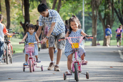 Full length of sisters riding bicycles with father on road at public park
