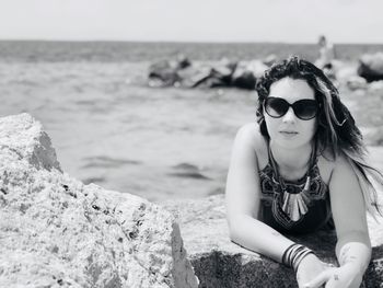 Young man wearing sunglasses on rock at beach against sky