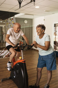 Cheerful senior woman in activewear laughing while standing near motivated husband exercising on cycling machine during workout in modern gym